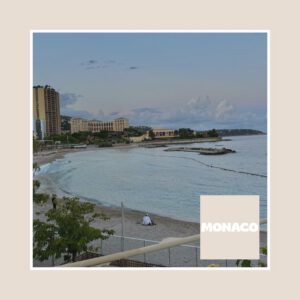 Photographie d’une plage de Monaco au coucher du soleil, avec une mer calme aux teintes bleu clair. À gauche, des bâtiments modernes, dont une tour haute, se dressent en arrière-plan. Quelques personnes se promènent ou sont assises sur le sable au premier plan. Le ciel présente des nuances pastel avec quelques nuages. En bas à droite de l’image, un carré beige avec le mot “MONACO” en lettres majuscules blanches ajoute une touche graphique.